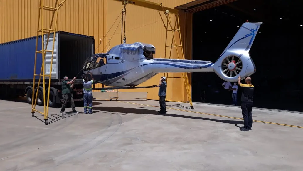 Helicopter suspended on a lifting frame being loaded into a shipping container inside a hangar