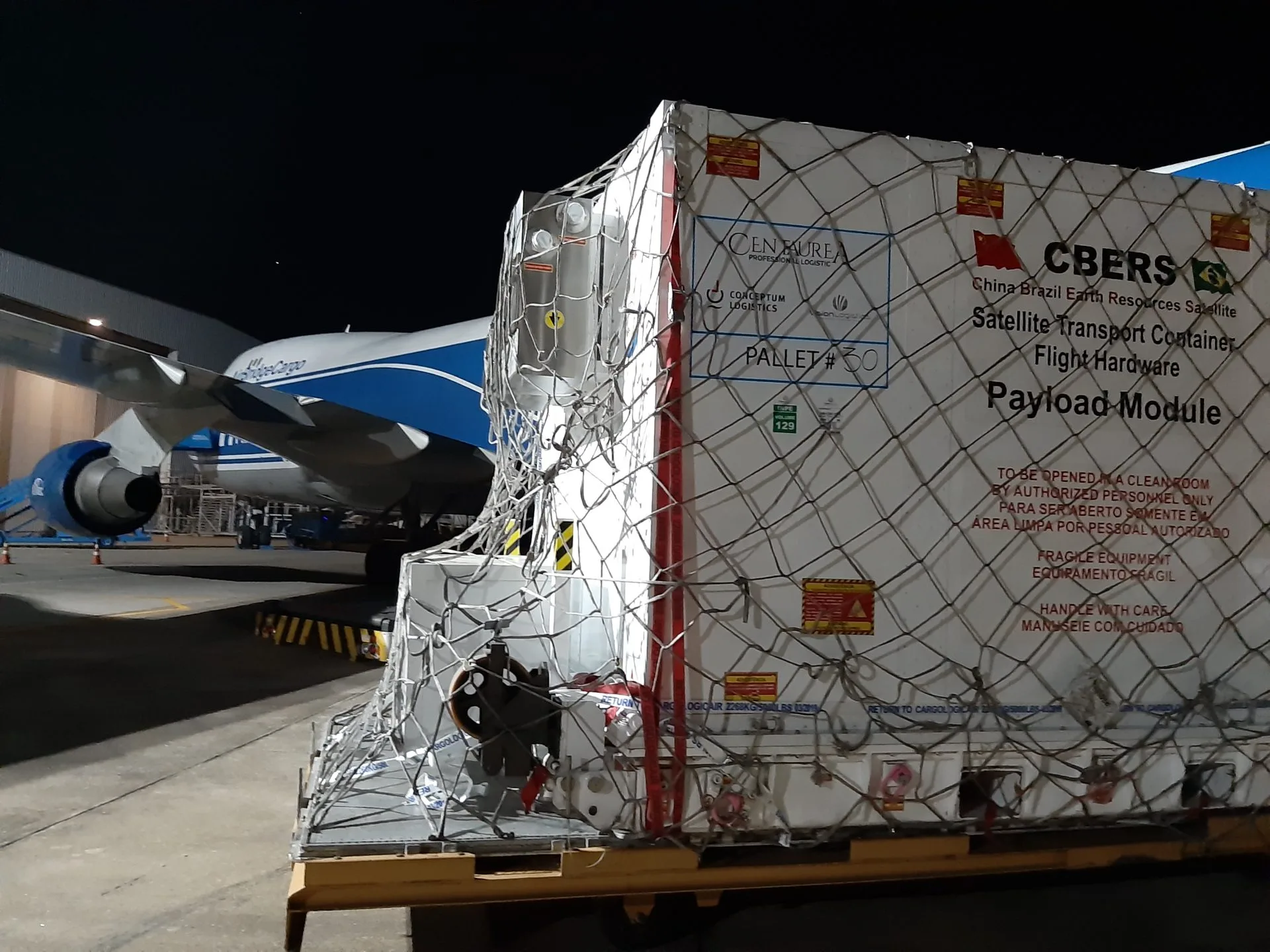 Satellite transport container loaded on a pallet beside a cargo aircraft at night during an air charter operation